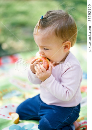 Little girl sits on a blanket and gnaws an apple. Close-up. Side view 88592199