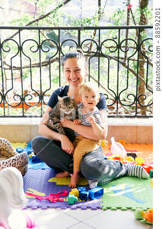 Mom sits on a rug on the balcony and holds a small child and a cat on her lap 88592201