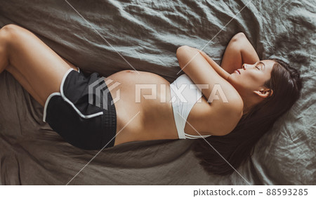 Pregnant woman sleeping lying on her side in bed during pregnancy. Top view of Asian model with first trimester belly in pajamas napping on top of sheets Pregnant woman sleeping lying on her side in bed during pregnancy. Top view of Asian model with first trimester belly in pajamas napping on top of sheets 88593285