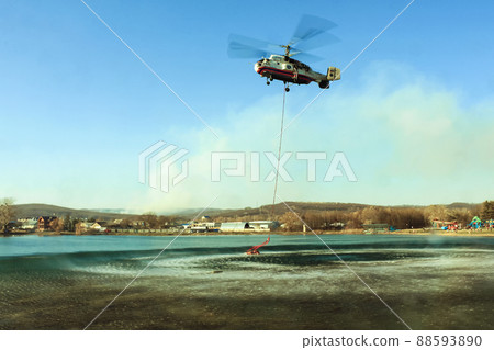A KA-32 helicopter of the Ministry of Emergency Situations hovering over a lake from which water is drawn to extinguish a forest fire. In the background is a blue sky. A KA-32 helicopter of the Ministry of Emergency Situations hovering over a lake from which water is drawn to extinguish a forest fire. In the background is a blue sky. 88593890