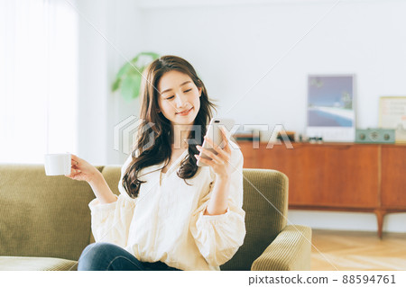 A woman sitting on the couch and looking at her smartphone while drinking coffee A woman sitting on the couch and looking at her smartphone while drinking coffee 88594761