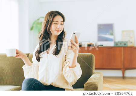 A woman sitting on the couch and looking at her smartphone while drinking coffee A woman sitting on the couch and looking at her smartphone while drinking coffee 88594762