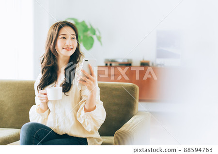 A woman sitting on the couch and looking at her smartphone while drinking coffee A woman sitting on the couch and looking at her smartphone while drinking coffee 88594763