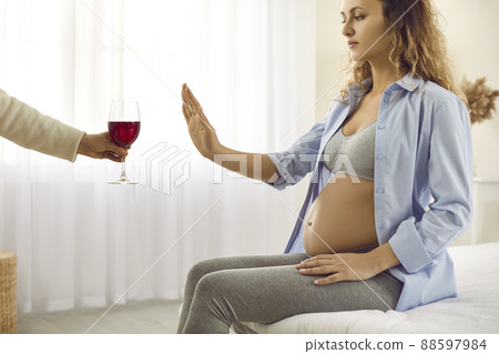 Pregnant woman being offered glass of wine doing stop gesture and refusing to drink 88597984