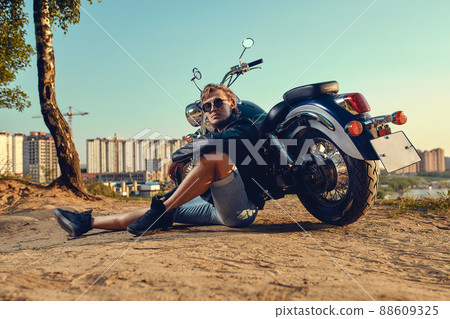 Sexy biker man wearing jeans and leather jacket sitting relaxed by his motorcycle 88609325