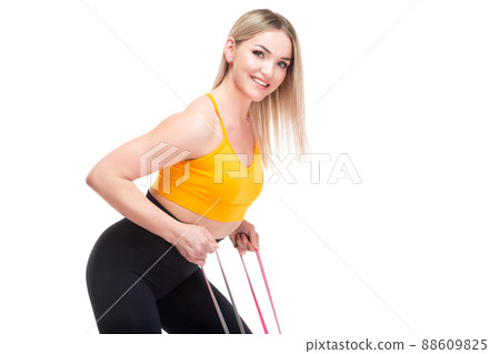 A beautiful, athletic, slim, smiling and cheerful woman in an orange top and black sweatpants performs an exercise with a red sports elastic band. Lifestyle concept with sports and gym, healthy 88609825