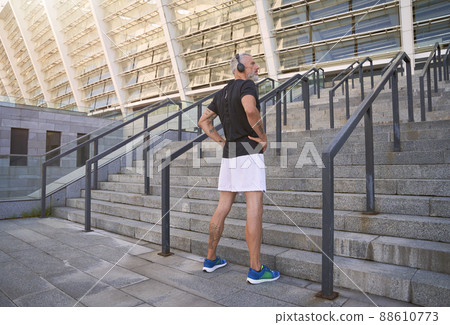 Full length shot of sportsman in headphones looking away while standing outdoors ready for morning workout in the city Full length shot of sportsman in headphones looking away while standing outdoors ready for morning workout in the city 88610773