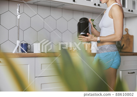Athletic woman shows large black jar of protein supplement shooting video in kitchen Athletic woman shows large black jar of protein supplement shooting video in kitchen 88610802