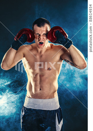 Winner of competitions. Studio portrait of a muscular boxer in professional gloves of European appearance with light bristles and hair on his chest. Smoke in background Winner of competitions. Studio portrait of a muscular boxer in professional gloves of European appearance with light bristles and hair on his chest. Smoke in background 88614934