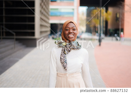 Positive well dressed African American female in Islamic headscarf standing in modern city district and looking towards the camera with wide smile 88627132