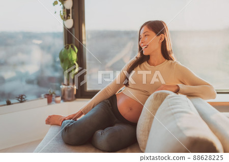 Happy Asian pregnant woman relaxing at home watching tv on sofa in living room. Laughing young girl during pregnancy Happy Asian pregnant woman relaxing at home watching tv on sofa in living room. Laughing young girl during pregnancy 88628225