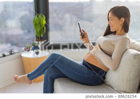 Pregnancy and maternity phone app for online prenatal class, preparation for birth. Happy Asian pregnant woman using cellphone at home watching videos on smartphone Pregnancy and maternity phone app for online prenatal class, preparation for birth. Happy Asian pregnant woman using cellphone at home watching videos on smartphone 88628254