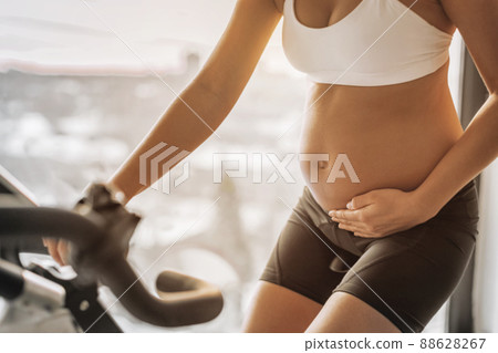 Pregnant woman training cardio on exercise spin bike at indoor fitness gym or home training on stationary bicycle. Closeup of fit girl holding pregnancy belly 88628267