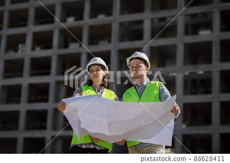 two engineers, a man and a woman in white helmets and protective vests, work at the construction site.  88628411