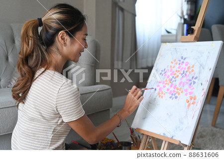 Smiling young asian woman sitting in front of easel and painting picture with water color on canvas. 88631606