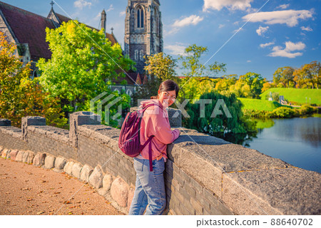 A beautiful young smiling brunette woman stands on a stone bridge near the St. Albans Church in the park Churchillparken. Copenhagen, Denmark A beautiful young smiling brunette woman stands on a stone bridge near the St. Albans Church in the park Churchillparken. Copenhagen, Denmark 88640702