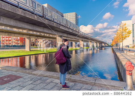A young woman stands near a canal with water and a bridge over which a subway metro train passes in the orestad city area in Copenhagen, Denmark 88644162