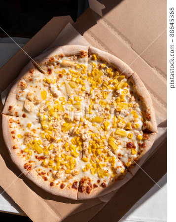 Tasty pizza with pineapple corn mozzarella cheese and white sauce in cardboard box on a table in daylight harsh shadows. Tasty pizza with pineapple corn mozzarella cheese and white sauce in cardboard box on a table in daylight harsh shadows. 88645368