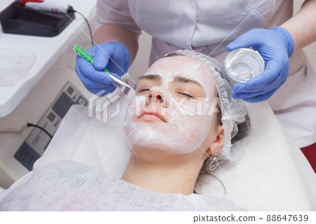 Master creates a white mask with a brush on the face of a young woman lying on the table. 88647639