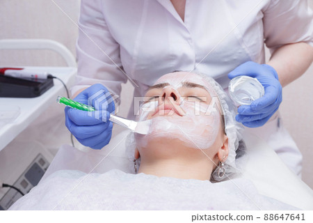 Crop master applying white mask with brush on face and neck of young woman in towel lying on table 88647641