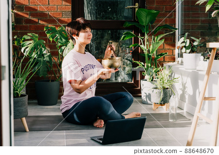 Young woman doing exercise yoga, playing on tibetan singing bowl with its mallet in cozy modern loft style room full of home plants on sunny morning. Online class yoga practice with meditation. 88648670