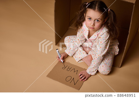 Top view little girl in pajamas writing No War on poster while sitting inside cardboard box, hiding from military and political conflict. Concept of refugees, immigrants losing their home during war 88648951