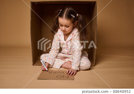 Beautiful child, adorable Caucasian kid girl hiding from the war inside a cardboard box and writing a social message Stop War. Concepts of refugees migrants losing their home during military conflict 88648952