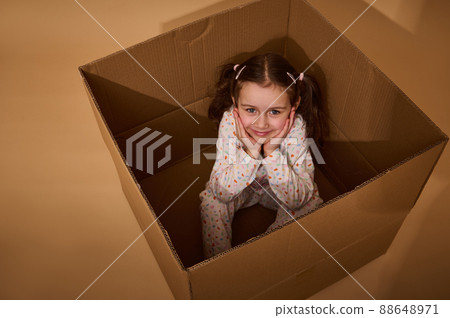 Top view of a charming little Caucasian girl sitting inside a cardboard box and looking at camera, isolated over beige background with copy space 88648971