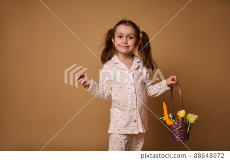 Portrait of a beautiful child girl in pajamas with bright dots stands with a purple basket and colored Easter eggs, isolated on a beige background with space for advertising text 88648972