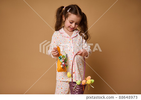 Cheerful adorable Caucasian baby girl in pajamas holding a rabbit toy and colorful decoration of Easter eggs, smiles posing to camera, isolated over beige background with copy ad space 88648973