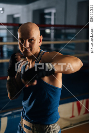 Vertical shot of bald aggressive boxer male wearing bandages punching air to camera in sport club with dark interior, on background of ring. Professional fighter fighting shadow looking at camera. 88651532