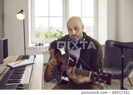 Songwriter and guitarist playing guitar while sitting by electronic keyboard in home studio 88652382