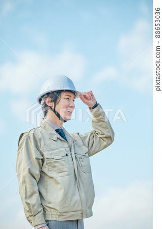 A young man in work clothes standing in a helmet against the blue sky 88653036