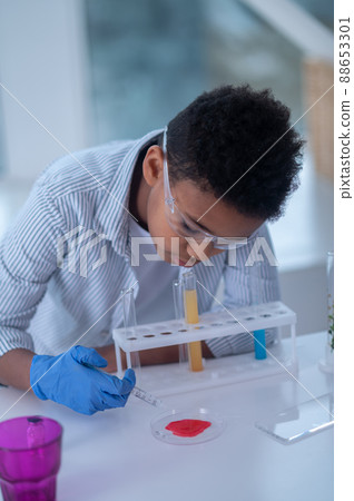 A dark-haired boy in a lab coat looking busy while doing chemical experiment A dark-haired boy in a lab coat looking busy while doing chemical experiment 88653301