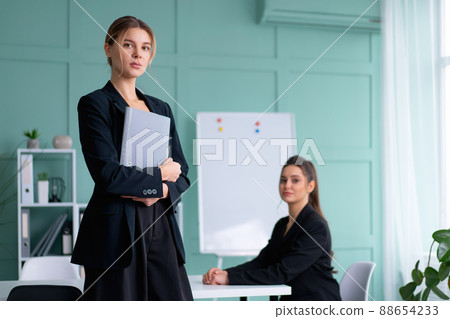 Two young women leaders dressed black suit in office one woman standing with document folder other woman sitting on the table looking at camera. Business meeting 88654233