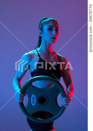 One muscled woman in sportswear training with disk of barbell isolated on purple background in neon light. Sport, beauty, strength concept 88676770