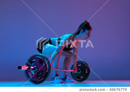 Portrait of muscled woman in sportswear training with a barbell isolated on purple background in neon light. Sport, weightlifting concept Portrait of muscled woman in sportswear training with a barbell isolated on purple background in neon light. Sport, weightlifting concept 88676779