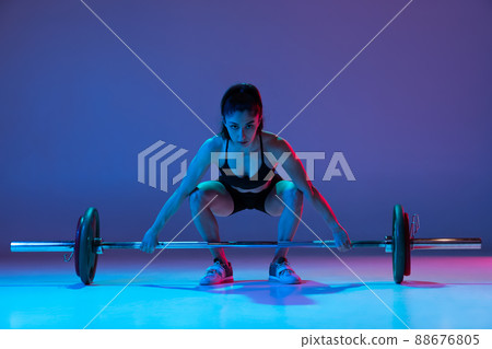Portrait of muscled woman in sportswear training with a barbell isolated on purple background in neon light. Sport, weightlifting concept Portrait of muscled woman in sportswear training with a barbell isolated on purple background in neon light. Sport, weightlifting concept 88676805
