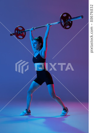 Portrait of muscled woman in sportswear training with a barbell isolated on purple background in neon light. Sport, weightlifting concept 88676832
