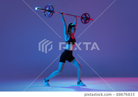 Portrait of muscled woman in sportswear training with a barbell isolated on purple background in neon light. Sport, weightlifting concept 88676833