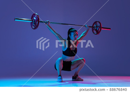 Portrait of muscled woman in sportswear training with a barbell isolated on purple background in neon light. Sport, weightlifting concept Portrait of muscled woman in sportswear training with a barbell isolated on purple background in neon light. Sport, weightlifting concept 88676840