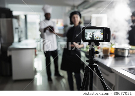 Close up of camera recording food industry workers preparing delicious gourmet dish while broadcasting. Chefs in restaurant professional kitchen filming cooking session for culinary school. Close up of camera recording food industry workers preparing delicious gourmet dish while broadcasting. Chefs in restaurant professional kitchen filming cooking session for culinary school. 88677031