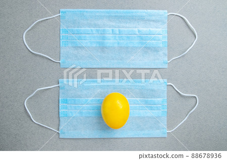 One yellow easter egg lies on a blue medical mask on a gray background. One yellow easter egg lies on a blue medical mask on a gray background. 88678936