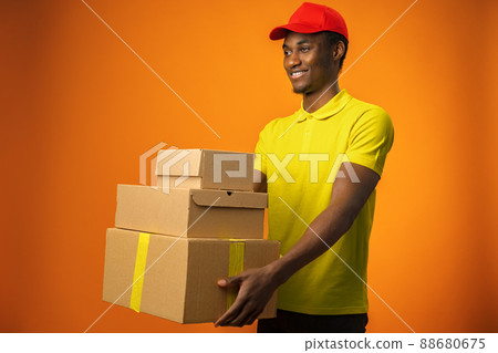 Smiling black male courier wearing uniform holding box in orange studio Smiling black male courier wearing uniform holding box in orange studio 88680675