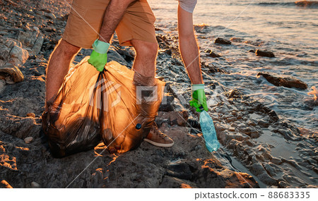 A male volunteer holding plastic bags with garbage and pick ups plastic bottle. Close up. The concept of coastal zone cleaning and world environment day 88683335