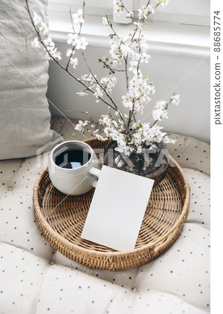 Spring still life composition. Greeting card mockup on wickered tray, cup of coffee. Feminine styled photo. Floral scene with blurred white cherry tree blossoms on bench near window. Selective focus 88685774