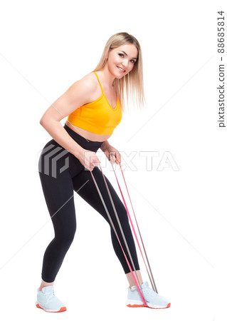 A beautiful, athletic, slim, smiling and cheerful woman in an orange top and black sweatpants performs an exercise with a red sports elastic band. Lifestyle concept with sports and gym, healthy A beautiful, athletic, slim, smiling and cheerful woman in an orange top and black sweatpants performs an exercise with a red sports elastic band. Lifestyle concept with sports and gym, healthy 88685814