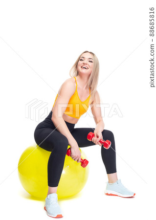 A beautiful, athletic, slim, smiling and cheerful woman in an orange top and black sweatpants performs exercises with dumbbells while sitting on a yellow fitball. Lifestyle concept with sports and gym 88685816