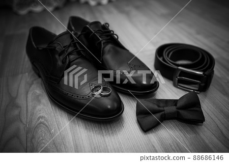 Leather black shoes, a belt, a pair of gold rings, a green butterfly tie on a brown background Leather black shoes, a belt, a pair of gold rings, a green butterfly tie on a brown background 88686146