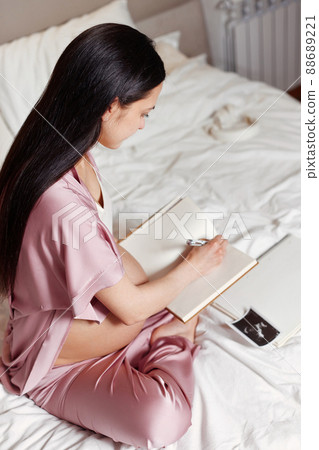 Pregnant woman sitting on bed and writing in notebook 88689221